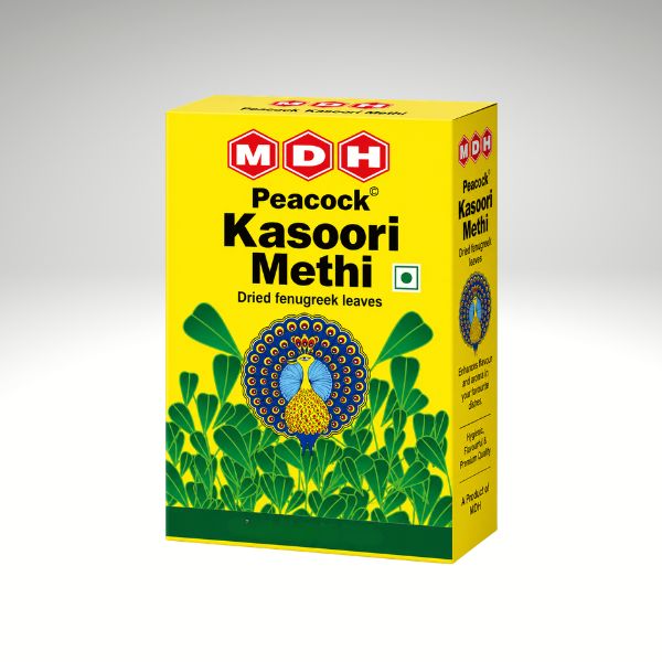 A yellow box of MDH Peacock Kasoori Methi (dried fenugreek leaves) against a neutral gray gradient background. The packaging features a bright yellow background, green leaf illustrations, a peacock logo, and the MDH brand logo in red and white.